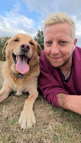 Blake, a yellow Labrador retriever 'smiling' with his tongue out as he lays on the grass next to his owner, Simon